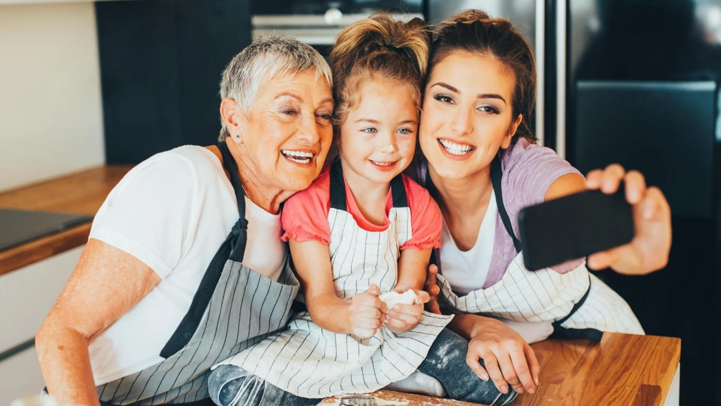 Nonna, mamma e bambina con il grembiule si scattano un selfie in cucina, davanti all’impasto